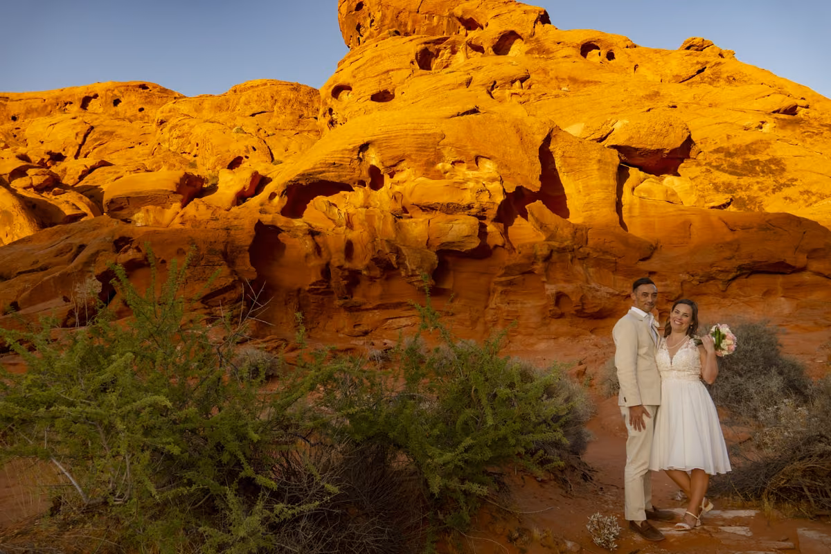 A bride and groom stand beneath towering golden sandstone cliffs at sunset in Valley of Fire, Nevada.