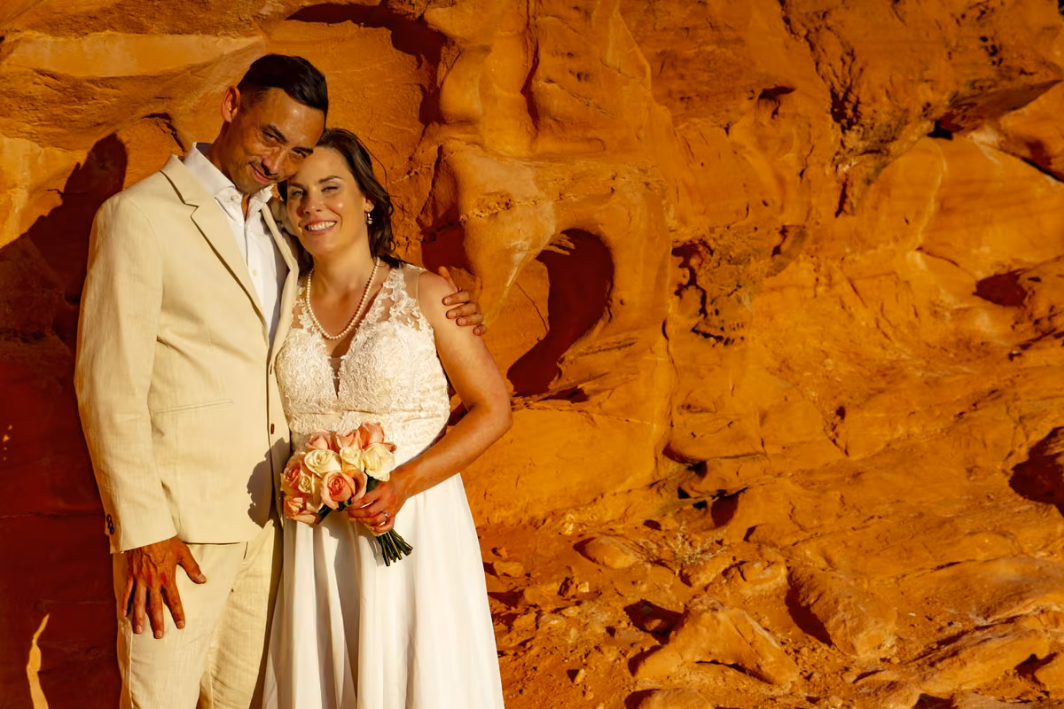 A bride and groom pose against a glowing orange sandstone wall at their Valley of Fire desert wedding.